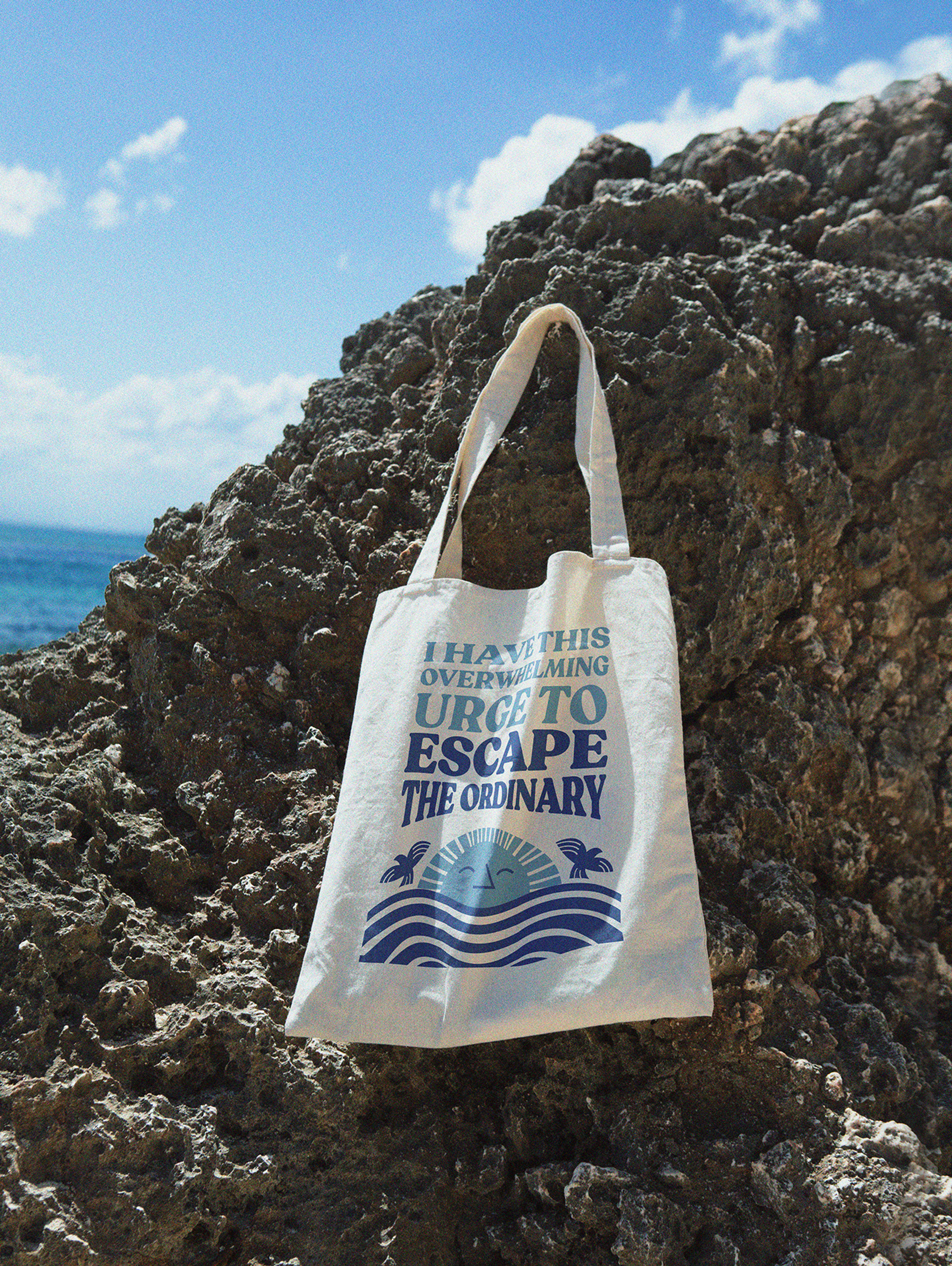 White Liberty Coast branded tote bag with slogan “I have this overwhelming urge to escape the ordinary” in front of coastal rocks designed by Our Revolution
